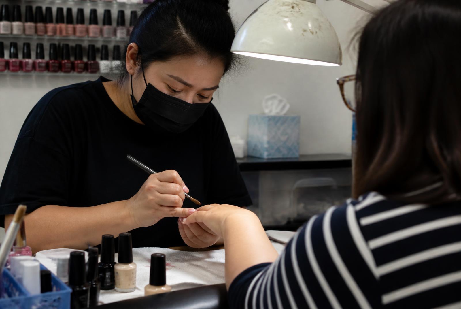 A nail technician finishing a gel manicure at a station under natural light, the client’s hand under the lamp